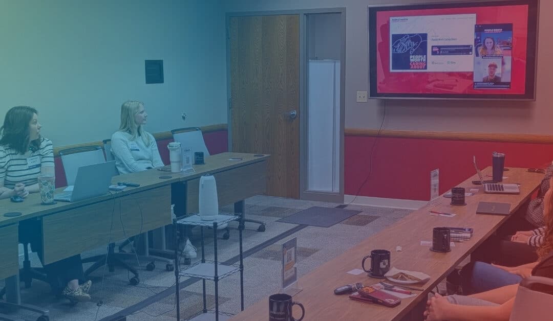 A group of people in a meeting room watch a presentation on a large screen.